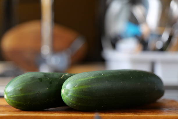 On the picture there are two cucumbers lying on a wooden chopping board in the kitchen of a home. In the background we see the sink and other things related to dishwashing.