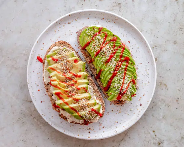 Two toasted bread slices topped with hummus, sliced avocado, sesame seeds and hemp seeds, with a drizzle of sriracha sauce