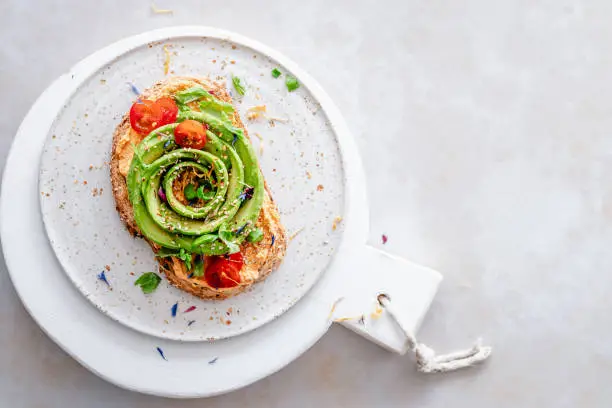 Overhead view of a toasted slice of bread with paprika hummus, avocado rose, cherry tomatoes, fresh basil, sesame seeds and edible flower petals, with copy space on the right side