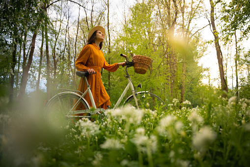 Young woman wearing yellow dress looking up while walking with bicycle amidst flowers in forest