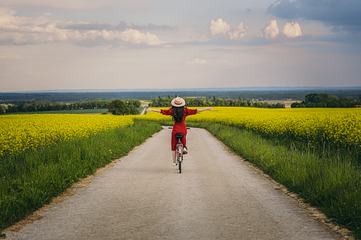 Rear view of joyful woman cycling with arms outstretched on road amidst canola farm against sky