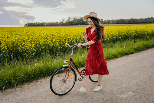 Smiling woman wearing red dress using cellphone while walking with bicycle on road against canola field