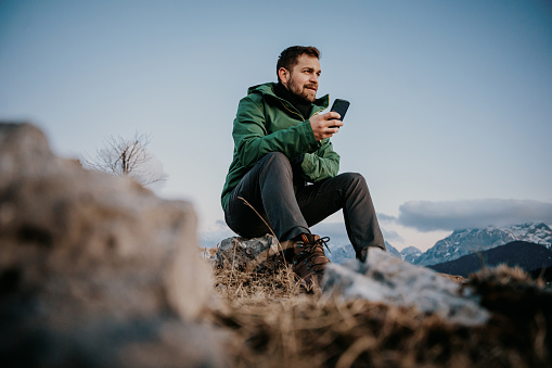 Male hiker text messaging on smart phone while sitting on stone at mountain top against blue sky