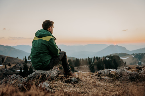 Male hiker enjoying beautiful view of sunrise while sitting on stone at mountain top against sky