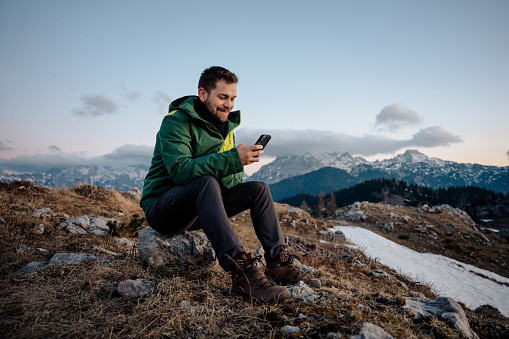 Smiling male hiker text messaging on smart phone while sitting on stone at mountain top against sky