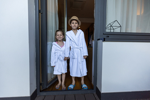 Brother and sister in white bathrobes holding hands and standing on hotel  terrace door