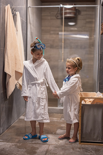 Little brother and sister in bathrobes  and swimming goggles standing and holding hands in bathroom