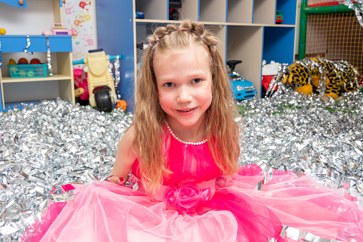 Smiling cheerful girl in a pink dress in a huge pile of silver foil confetti in the nursery. A girl in sparkling sequins. Happy children's birthday, Christmas, New Year. The concept of the holiday.