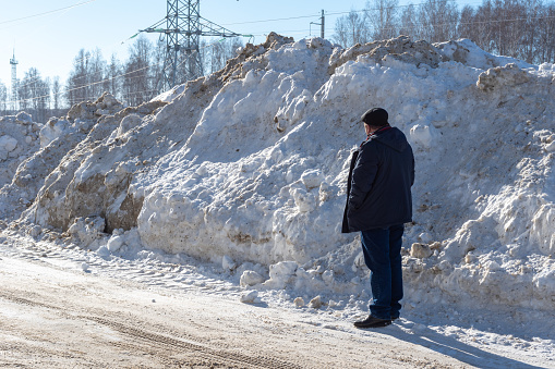 A man standing next to a large pile of snow on the side of the road. High snowdrifts after a snowfall or blizzard. Clearing snow on roads for safe driving