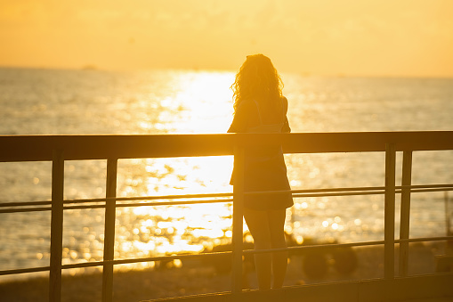 silhouette of a beautiful girl who stands on the bridge in the rays of the setting sun and takes pictures on the phone as the sun sets over the sea in the evening, selective focus