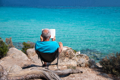 Portrait of a handsome man lying on a deckchair and reading a book. Happy mature man next to  the beach on Aegean sea with beautiful view. Summer vacation concept.