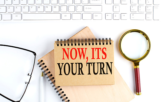 NOW,IT'S YOUR TURN text in office notebook with keyboard, magnifier and glasses , business concept
