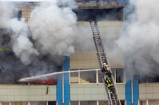 Firemen on telescopic ladder extinguishing strong fire with from a fire fighting firehose nozzle in rescue operation.Thick grey smoke and fire from the windows without glass of industrial warehouse.