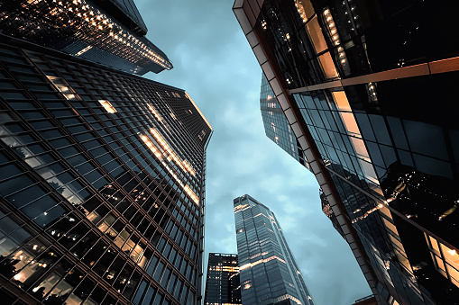 City of London at night, looking up at tall skyscrapers against a dramatic sky. Office lights illuminate the dark building surfaces revealing the interiors. Glass and steel geometries with reflections. Modern architecture photography.