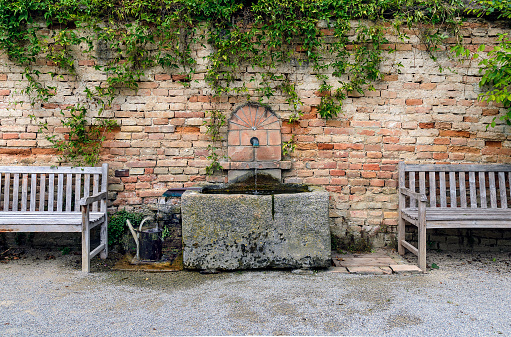 wall fountain with stone trough between two wooden settees before a brick wall at palace Schlosshof in Austria