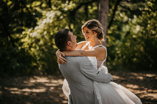 Young cheerful groom carrying and spinning his new wife in nature on a wedding day