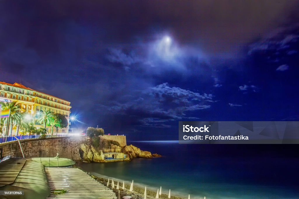 Fantastic night view on amazing Mediterranean Sea and part of building ( maybe Hotel)  with Moon Fantastic night view on amazing Mediterranean Sea and part of building ( maybe SwissNice Hotel)  with Moon and clouds   and Rob Kape promenade -  and folded umbrellas from the sun on the beach - French Riviera! Beach Stock Photo