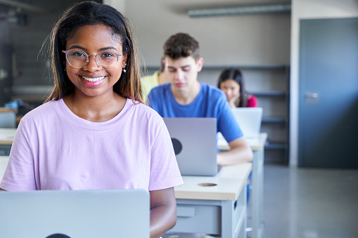 Smiley portrait of African american young woman looking at camera while studying at the school, using a laptop with her classmates on the background. High quality photo