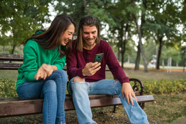 istock Two friends having fun time together watching videos using smart phone at public park 1413120483