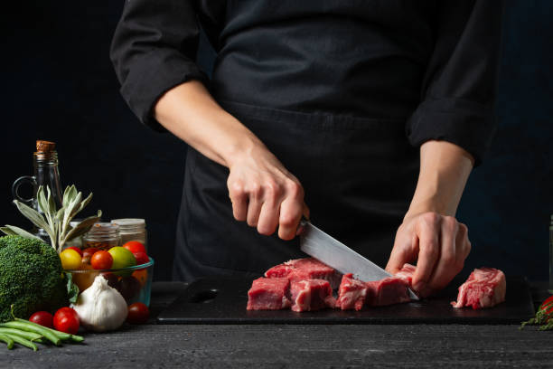 プロのシェフがダークブルーの背景に黒板に新鮮な牛肉をナイフでカット。リブアイステーキの準備の舞台裏。クローズアップ ビュー。食品コンセプト。 - rib eye steak beef cutting board meat ストックフォトと画像