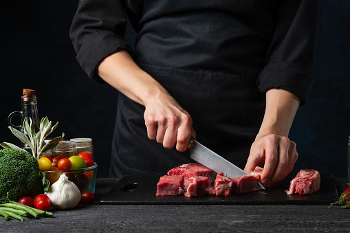 The professional chef cutting with knife fresh beef meat on the black board on dark blue background. Backstage of preparing rib-eye steak. Close-up view. Food concept.
