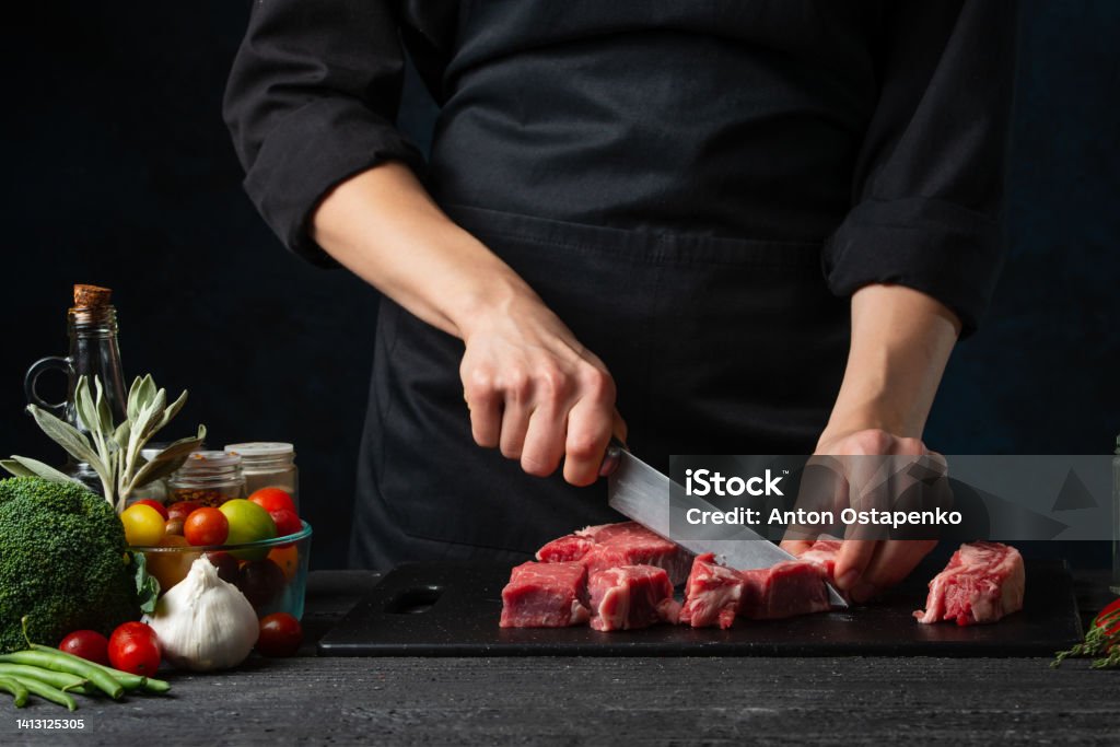 The professional chef cutting with knife fresh beef meat on the black board on dark blue background. Backstage of preparing rib-eye steak. Close-up view. Food concept. Meat Stock Photo