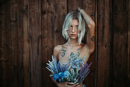 Blonde woman posing with tattoos all over her body in front of a wooden wall. Colourful and styled outfit.