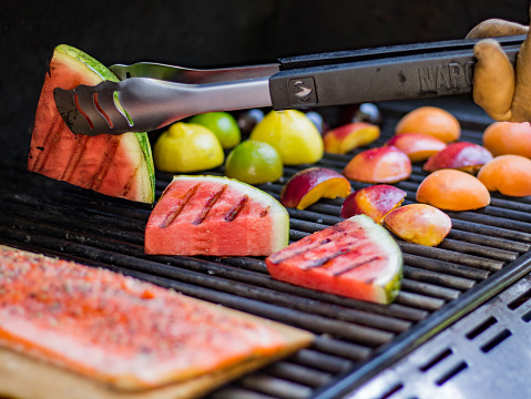 A very healthy summer meal of grilled watermelon, peaches, nectarine, plums, lemon and lime with a salmon fillet being cooked on a cedar plank. Hand with tongs turning watermelon over.