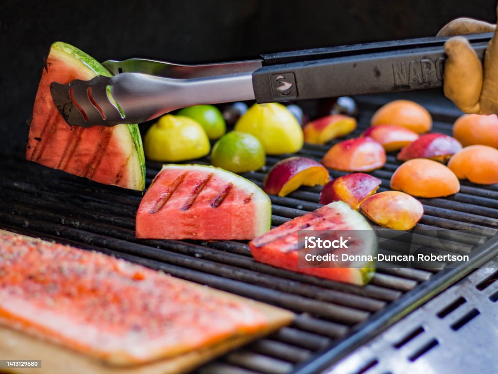 A summer barbecue  cooking up grilled fruit and salmon fillet on a plank for a fresh meal. A very healthy summer meal of grilled watermelon, peaches, nectarine, plums, lemon and lime with a salmon fillet being cooked on a cedar plank. Hand with tongs turning watermelon over. Grilled Stock Photo