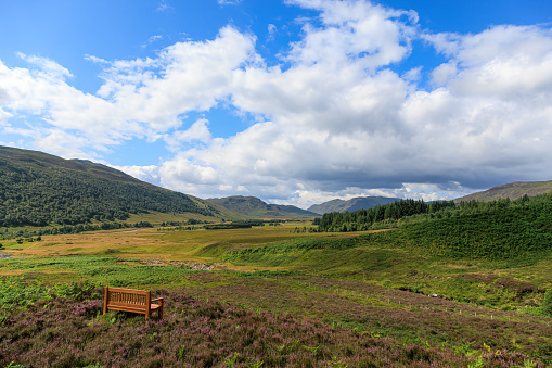 A scenic view of wooden bench facing a majestic Scottish mountain valley under a beautiful blue sky and some white clouds