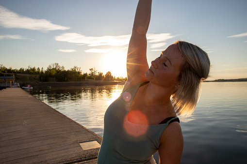 A woman is doing yoga by a lake in the sundown. She has a green suit. He is doing a yoga pose. 
you can see the sunbeams.