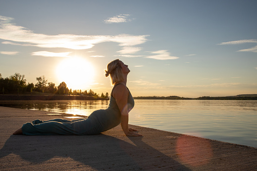 A woman is doing yoga by a lake in the sundown. She has a green suit. He is doing a yoga pose. 
you can see the sunbeams.