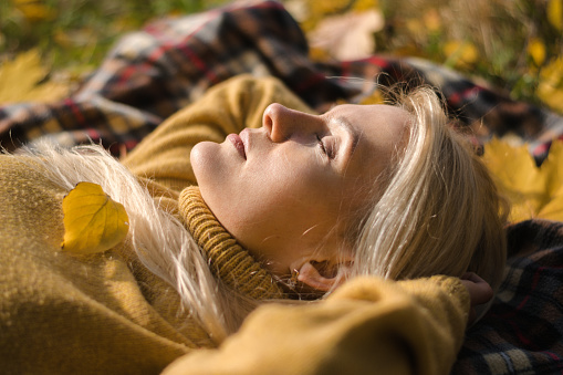 Attractive blonde woman in a yellow sweater lies on a blanket in the autumn foliage in the park.Autumn concept.Beauty in nature.Slow living.