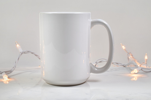 A blank 15 ounce coffee mug rests merrily atop a white marble background. Glowing white Christmas lights grace the background of this coffee cup mockup.