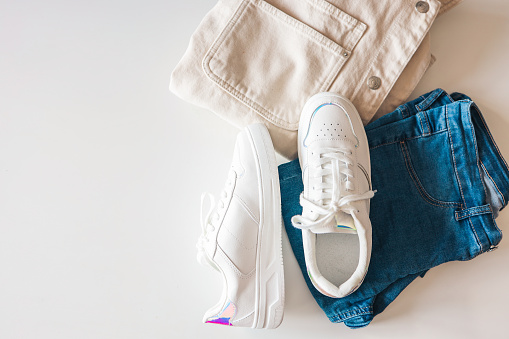 Stylish women's clothing flat lay. White sneakers, jeans and a jacket on a white background top view.