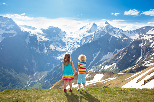 Children hiking in Alps mountains. Kids look at snow covered mountain in Austria. Spring family vacation. Little boy and girl on hike trail in blooming alpine meadow. Outdoor fun and healthy activity.