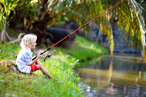 Boy fishing. Child with red rod catching fish in river on sunny summer day. Outdoor and nature activity for kids. Little boy sitting at lake shore. Children catch fish. Camping fun.