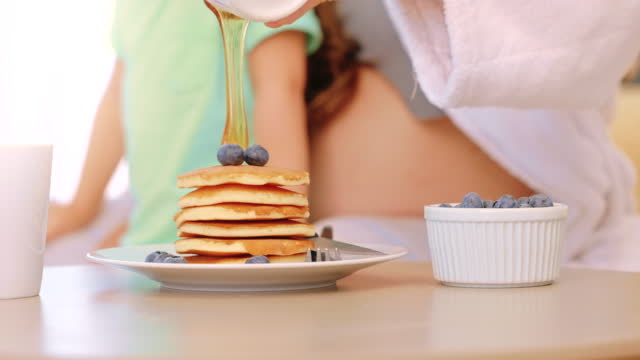 Syrup and pancakes for breakfast in the morning. Closeup of honey being poured over a stack of flapjacks at home on the weekend. A hungry single mother and her child eating fresh food in the bedroom