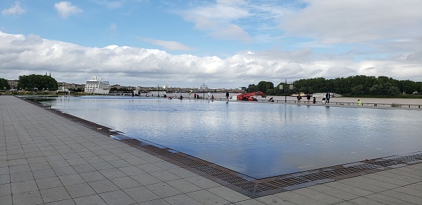 A panoramic view of the water mirror in Bordeaux, France. The mirror is the reflective nature of ghebthin film of water that sits on a very large and wide area of floor tiles, situated outside. The mirror reflects the sky and passing clouds.