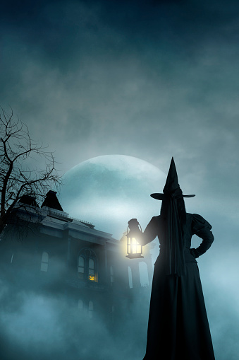 A rear view of a Halloween witch as she holds a lantern and looks up at an illuminated jack o'lantern sitting in the window of an old haunted house that is shrouded in fog and mist and silhouetted by a large rising moon on a dark and spooky Halloween night