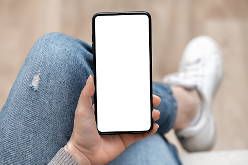 Top view mockup image of woman holding mobile phone with blank white screen. woman using cellphone with while sitting cross-legged on sofa at home. Mockup smartphone