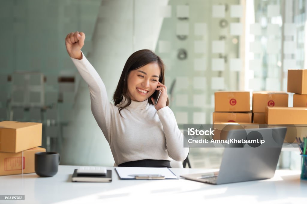 Young Asian small business owner successful selling online and holding smartphone with laptop computer. Happy Asian business woman successful excited raised hands rejoicing. Winning Stock Photo