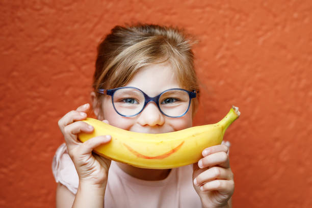 オレンジ色の背景に笑顔のような黄色いバナナを持つ幸せな小さな子供の女の子。眼鏡をかけた未就学児の女の子が微笑んでいる。子供のための健康的な果物 - バナナ ストックフォトと画像