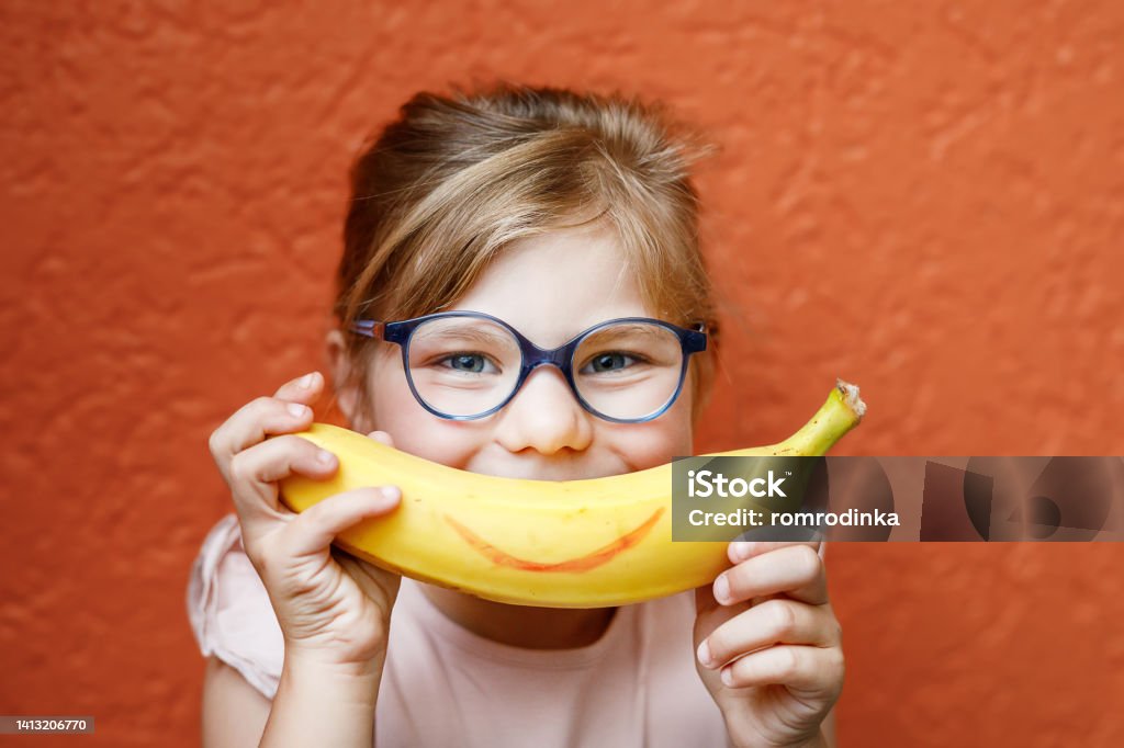 Happy little child girl with yellow banana like smile on orange background. Preschool girl with glasses smiling. Healthy fruits for children Happy little child girl with yellow banana like smile on orange background. Preschool girl with glasses smiling. Healthy fruits for children. Child Stock Photo