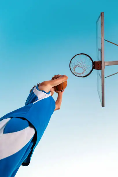 Photo of Basketball. A teenager in blue sportswear throws a ball into a basketball Hoop. Bottom view. Copy space. Sky in the background. Concept of sports games