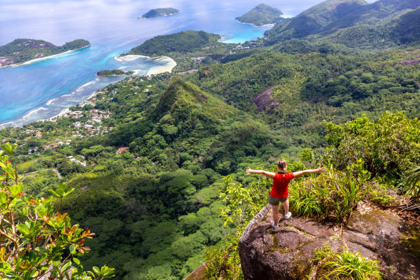 Young female traveler standing at the edge of the cliff at Morne Blanc View Point, Young female traveler standing at the edge of the cliff at Morne Blanc View Point, overlooking Mahe Island coastline with lush tropical vegetation and crystal blue ocean, Seychelles. mahe island stock pictures, royalty-free photos & images