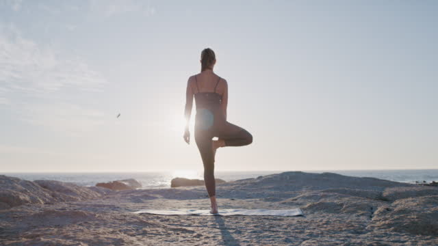 Healthy, fit and active young woman doing yoga on the beach with a beautiful view of the sea and horizon at sunset. Calm, peaceful and relaxed female yogi meditating for mental health and wellness