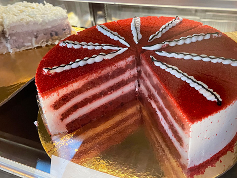 Stock photo showing close-up view of bakery display cabinet of an indulgent dessert of sliced red velvet cake, topped with piped icing, on circular, silver cake board available for purchasing by the slice.
