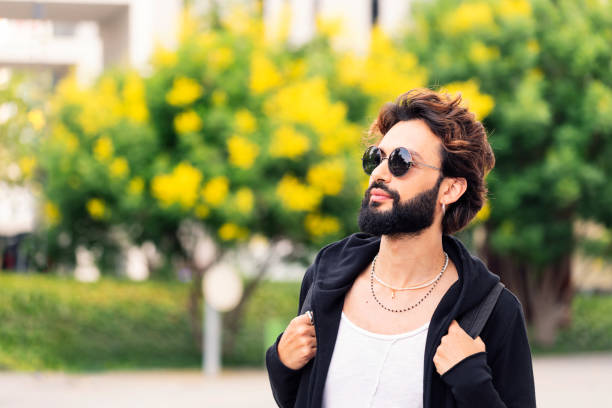 portrait of a stylish young college student with beard and sunglasses walking happy by the university campus, concept of youth and urban life style, copy space for text