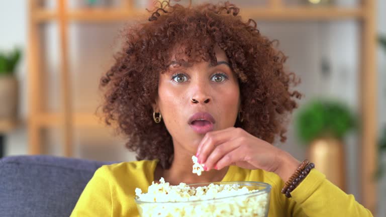 Woman watching a movie and eating popcorn while relaxing at home. Shocked female entertained by scary horror film and enjoying her movie streaming subscription with a variety of shows to watch